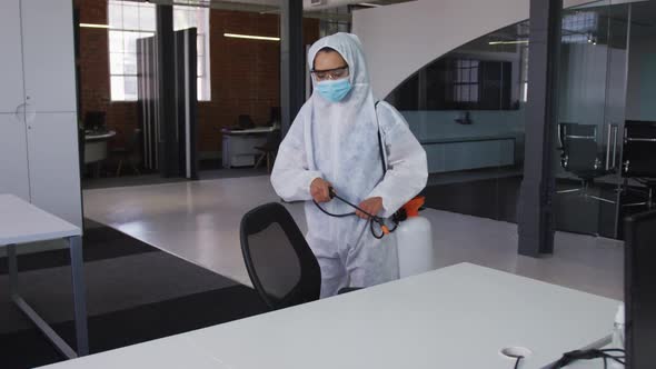 Female cleaner wearing protective face mask and overalls disinfecting office and furniture