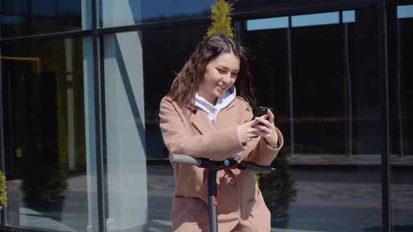 A Young Student Girl in a Sweater Jeans and a Coat with an Electric Scooter Stands Near a Stylish