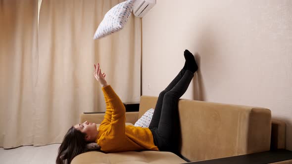 Bored Young Woman in Sweater Lies on Sofa Throwing Pillows