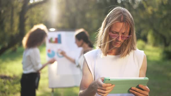 Portrait of Thoughtful Concentrated Young Man Surfing Internet on Tablet with Blurred Women