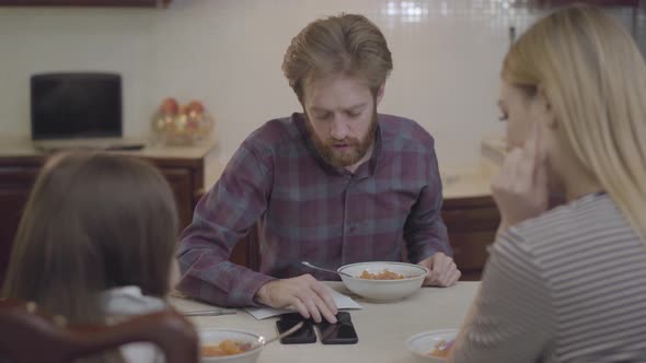 The Family of Three People Having Breakfast Sitting at the Table. Busy Father Talking By Cell Phone