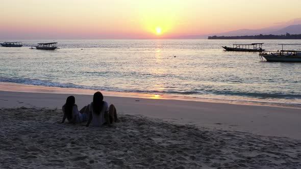 Young women sitting on sandy exotic beach, watching beautiful sunset reflecting sunlight on calm sea