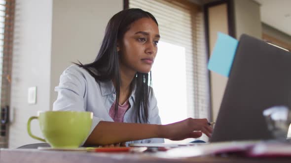 Mixed race woman using laptop writing in notebook drinking coffee working from home