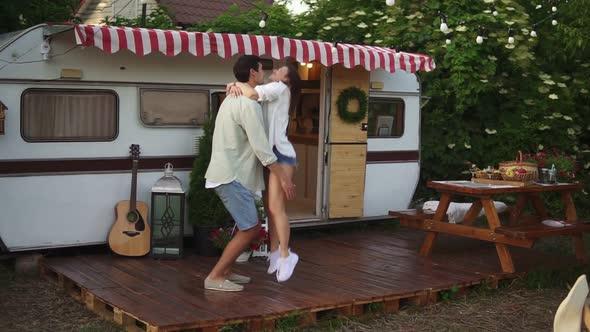 Happy Young Lovely Couple Having Fun Near Trailer in the Park Girl Jumping on Man's Arms He's