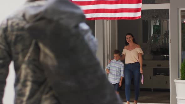 Caucasian male soldier greeting son and wife in garden with american flag hanging outside house