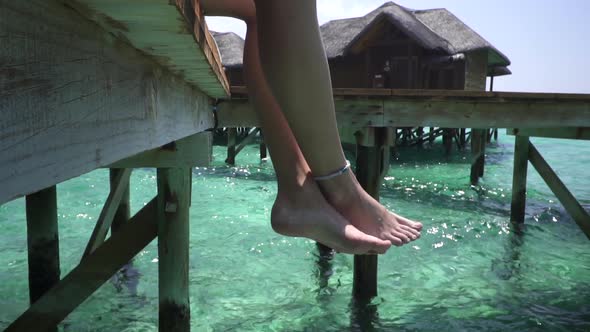 Young lady sitting on wooden jetty. Crossed legs swinging above turquoise ocean. Maldives. Bungalow