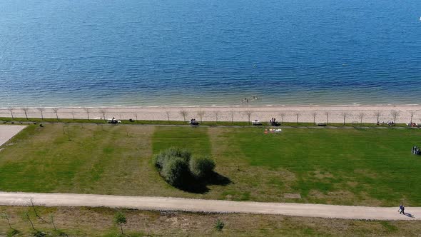 Lake beach with people walking and sunbathing and bathing, lawn garden with shrubs. Overhead drone s