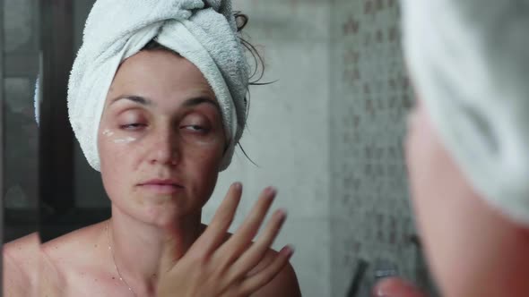 Young Woman is Applying Cream on the Face Eye Area in Bathroom Looking at Mirror