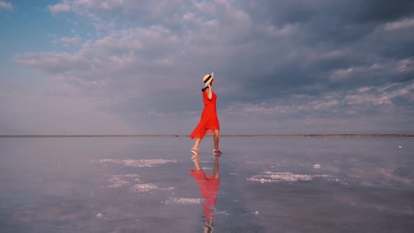 Hiking Girl in a Fluttering Dress Walks Along a Pink Salt Lake in Which the Sky Is Reflected