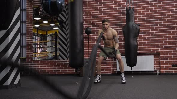 A Muscular Male Boxer Does an Exercise with Ropes in a Sports Gym