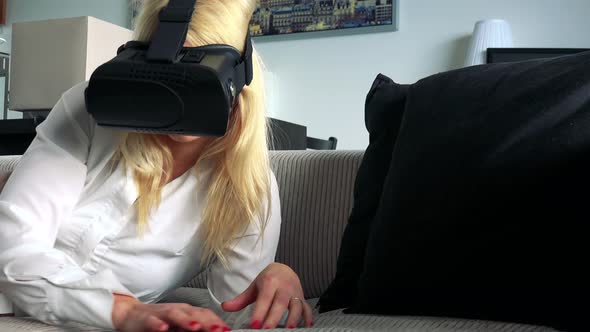 Young Attractive Blond Woman Lies on the Couch in Living Room and Uses Virtual Reality Glasses