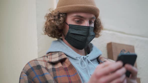 Concentrated curly-haired man in protective mask texting by phone