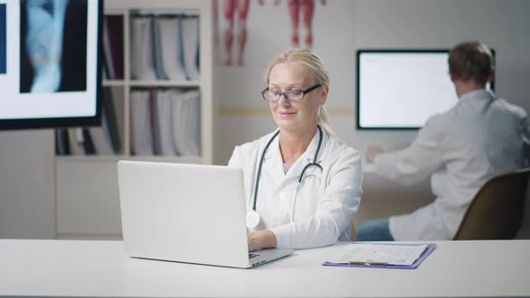 Portrait of Mature Woman Doctor Working on Laptop and Writing in Patient History