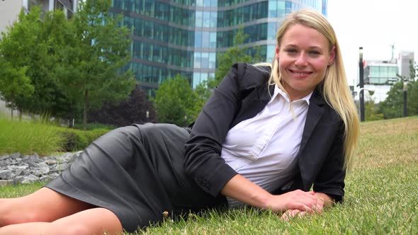 A Young Beautiful Businesswoman Lies in Grass in a Park and Smiles at the Camera - Office Buildings