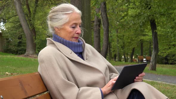 An Elderly Woman Sits on a Bench in a Park and Works on a Tablet
