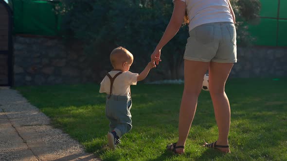 Mother Holds Hand of Little Son Hitting Soccer Ball on Grass