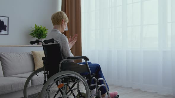 Middle Aged Woman in Wheelchair Looking at Window Alone Warming Her Hands and Riding Away at Home