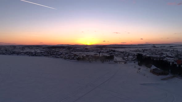 Aerial Over the Frozen Winding River Surrounded with Forest and with a Little Village on a Side