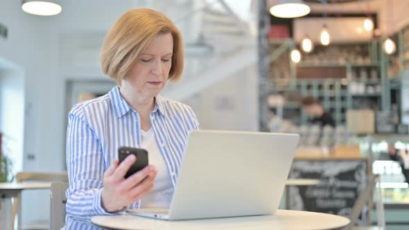 Smartphone Use By Creative Old Woman with Laptop in Cafe 