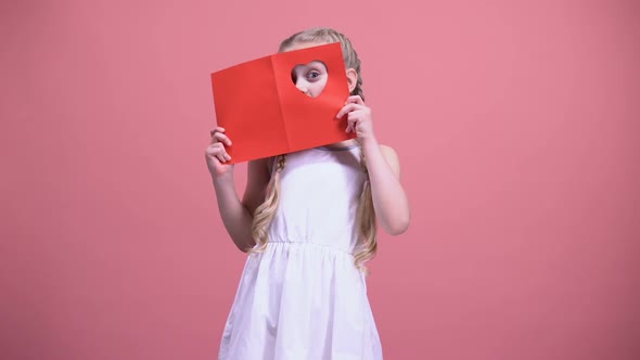 Little Girl With Braids Looking Through Heart-Shaped Hole in Valentine Card