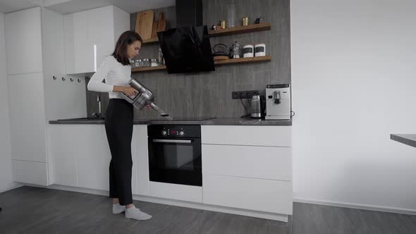 Slender Brunette Vacuums in the Kitchen. A Modern Wireless Vacuum Cleaner.