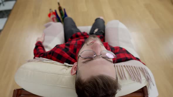 Top View of Drunken Young Man in Eyeglasses Leaning on Armchair with Blurred Alcohol Bottles