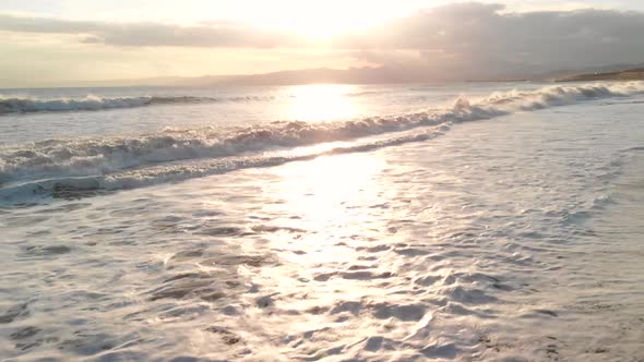 Aerial upward flight over waves crashing at beach at sunset while getting close to water level