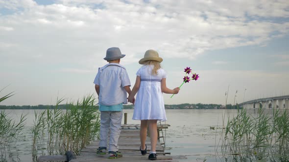Happy Sweet Children Boy and Girl with Toy Windmills Walk By Hand on Wooden Bridge on River Between