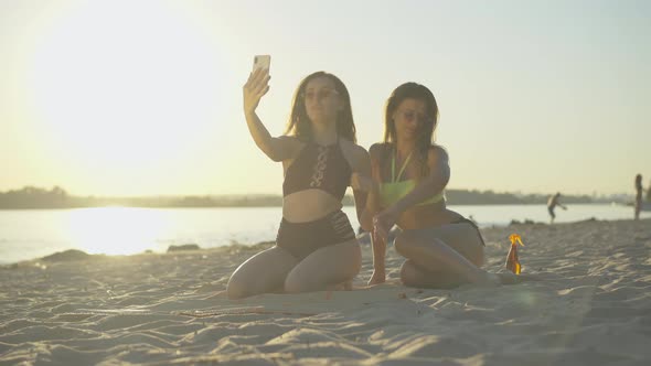 Wide Shot of Two Beautiful Female Tourists Sitting on Sandy Beach at Sunset and Chatting Online