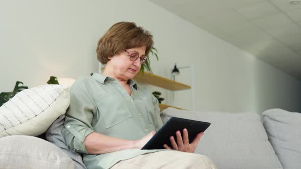 Smiling Elderly Woman Resting on Sofa Using Digital Tablet at Home