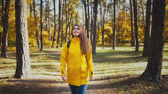 Young Happy Woman in Parka Jacket and Backpack is Smiling and Looking Around Walking By a Pathway in