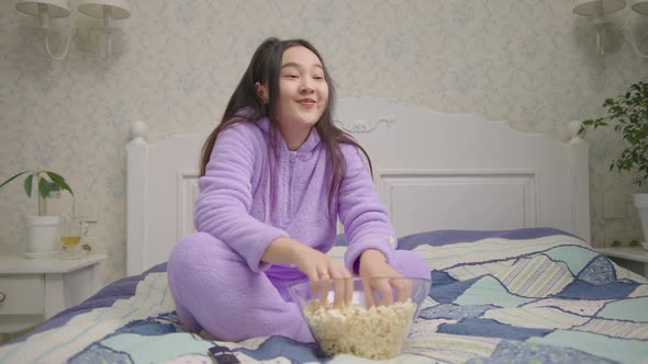 Asian Woman Watching Funny Movie or Tv Show and Eating Popcorn Sitting Alone on Bed