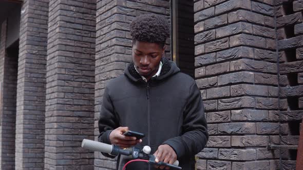 A Young African American in Casual Dark Clothes with a Scooter Stands Near a Building with a Dark