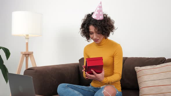 Happy Woman in Party Hat Opens Gift Box and Surprised Sitting in Couch at Home