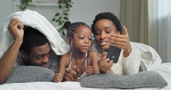 Afro American Family Black Ethnic African Parents Lie on Sofa Bed Covered with White Blanket Look at