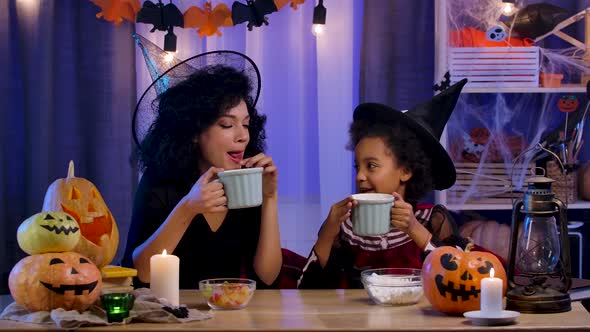 Mom and Daughter African American in Festive Costume and Witch Hat Having Fun and Drinking Hot