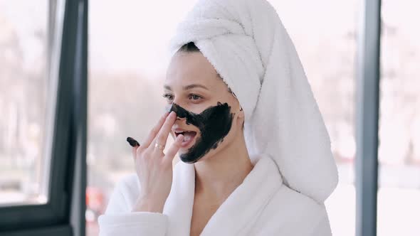 Pretty Woman with Wrapped Hair in a Towel Is Applying Black Mask on Face