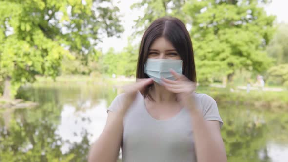 A Young Caucasian Woman in a Face Mask Celebrates in a Park  a Pond and Trees in the Background