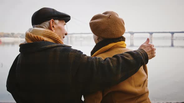 Aged Husband Hugging His Wife Sitting on Bench at Bank of a River Looking at Picturesque Riverscape