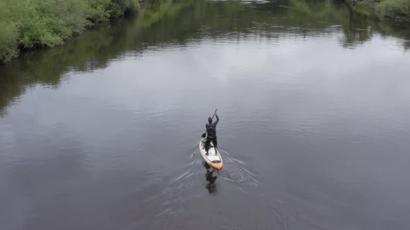 Aerial follows stand up paddleboarder paddling on small rural river