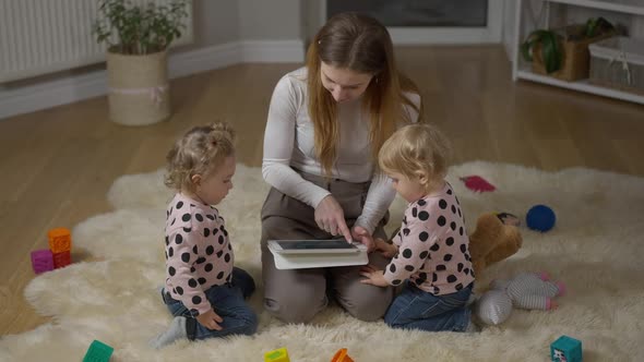 Wide Shot of Happy Millennial Woman and Twin Girls Sitting on Floor in Living Room Using Tablet