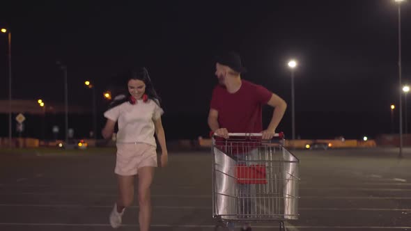 Two Cheerful Young People Running with Trolley Along Parking Lot at Dusk. Portrait of Happy Carefree