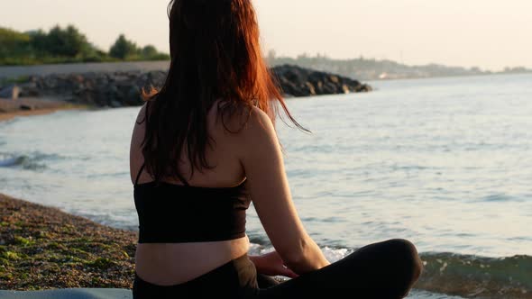 A yogi woman sits in a lotus position on the seashore and looks at the sea dawn of the sun