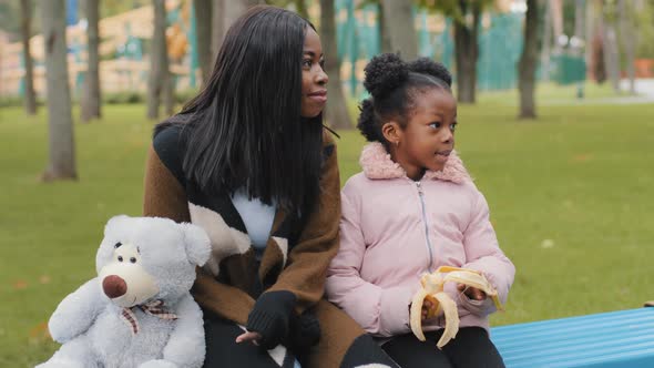 Portrait Young Mom and Daughter Sitting on Bench with Teddy Bear Child Eating Banana Woman Points