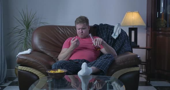 Portrait of Plump Redhead Caucasian Man Sitting in Front of TV and Drinking Beer. Lonely Single Guy