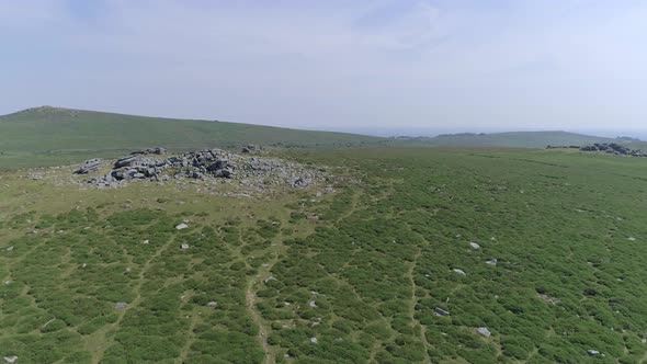 Forward tracking aerial towards a Dartmoor tor or rocky outcrop surrounded by green summer moorland.