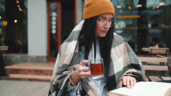 Smiling young brunette woman wearing hat and eyeglasses reading book and drinking tea