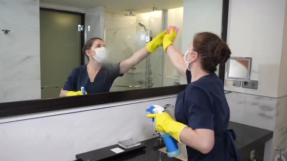 Masked Maid Spraying and Wiping Bathroom Mirror