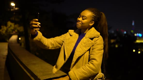 A Young Black Woman Takes Selfies with a Smartphone As She Sits in a Park in an Urban Area at Night