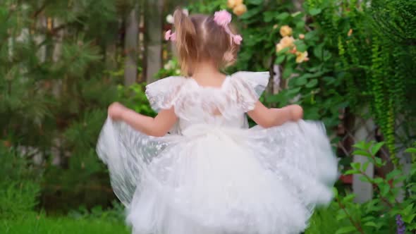 Happy Little Girl in a Smart White Dress Runs on the Lawn in the Garden